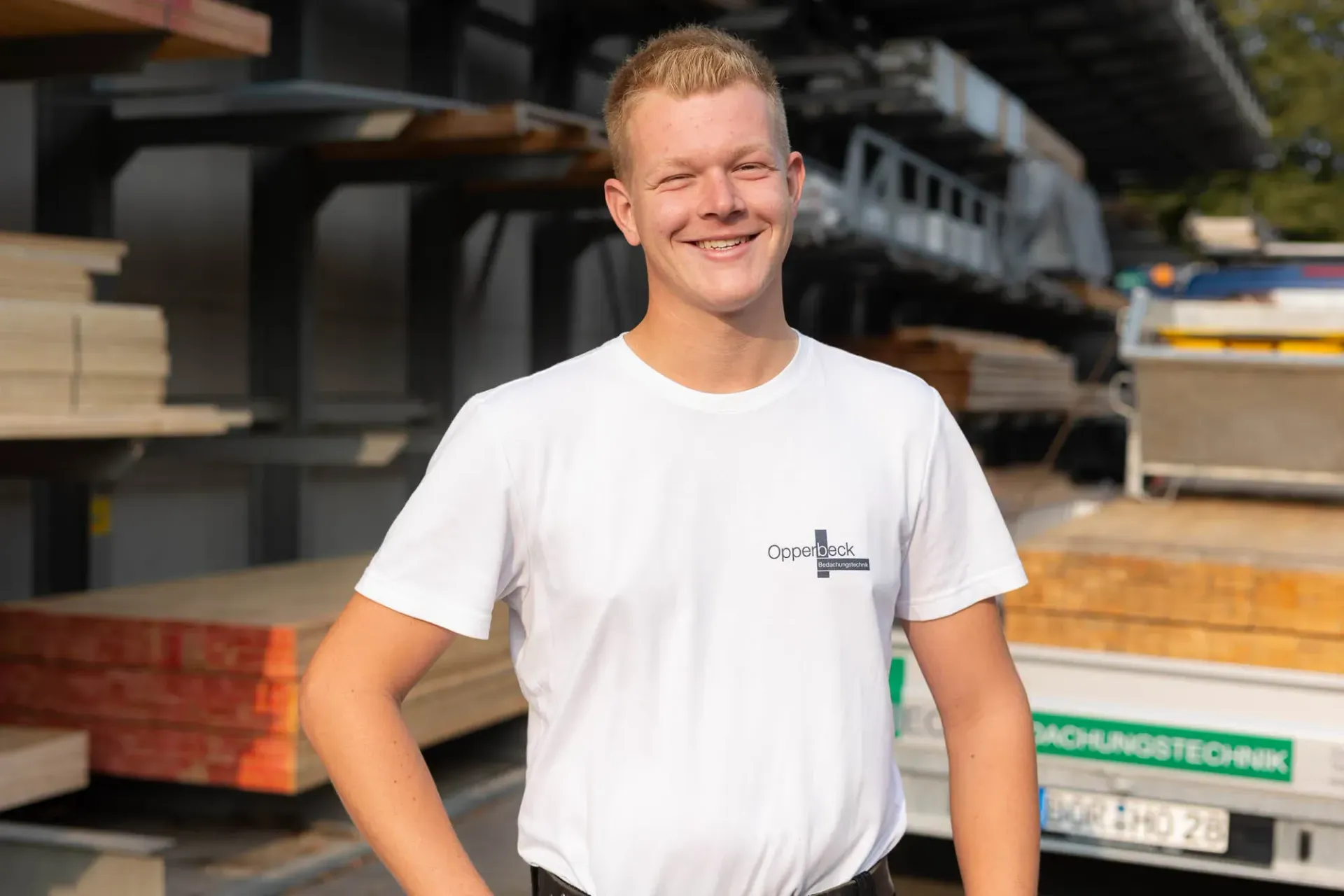 Ein junger Mann in einem weißen T-Shirt steht vor einem Holzstapel.