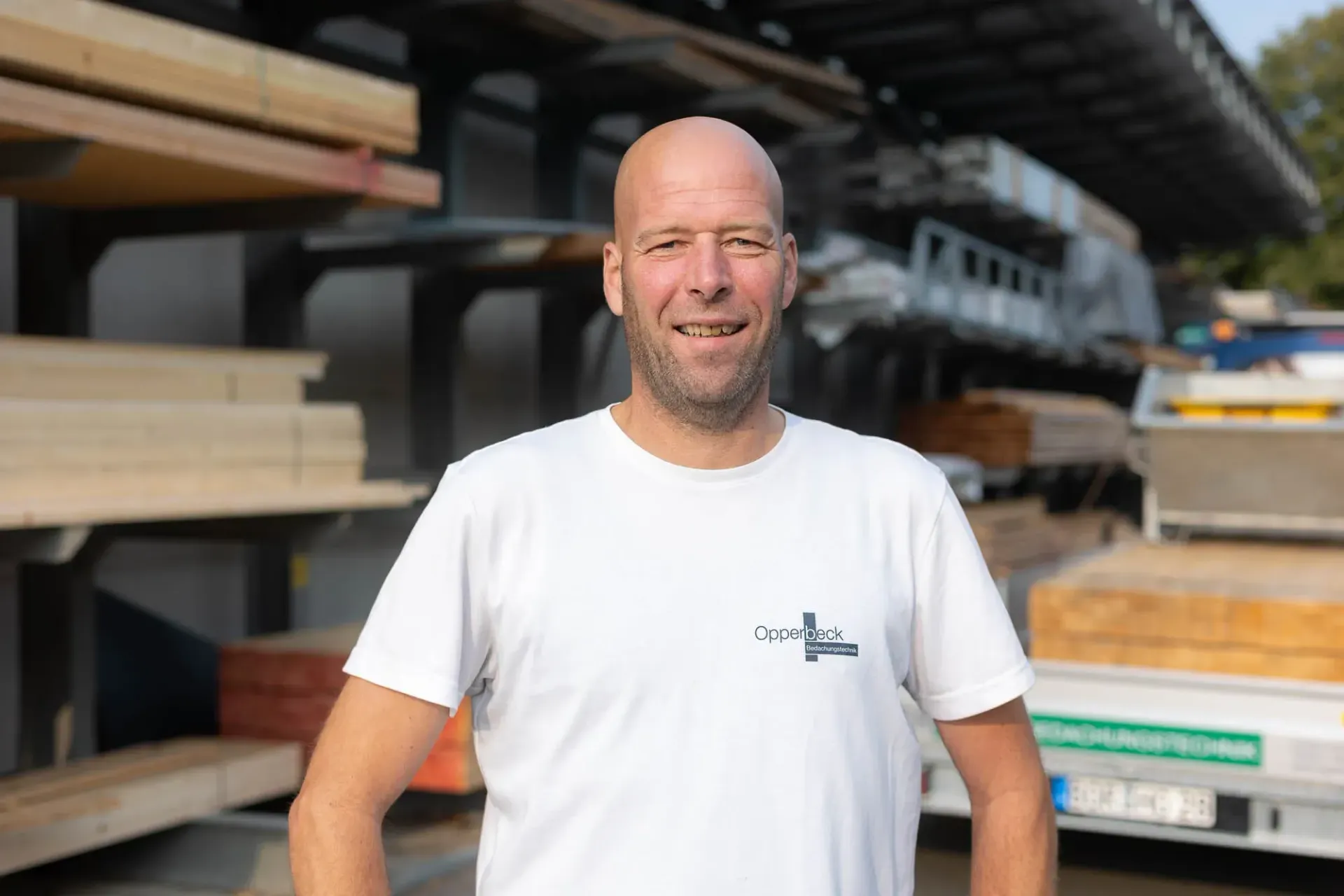Ein Mann im weißen Hemd steht vor einem Holzstapel.