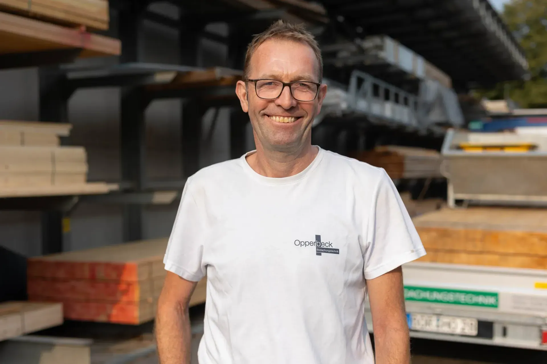Ein Mann mit Brille und weißem T-Shirt steht vor einem Holzstapel.
