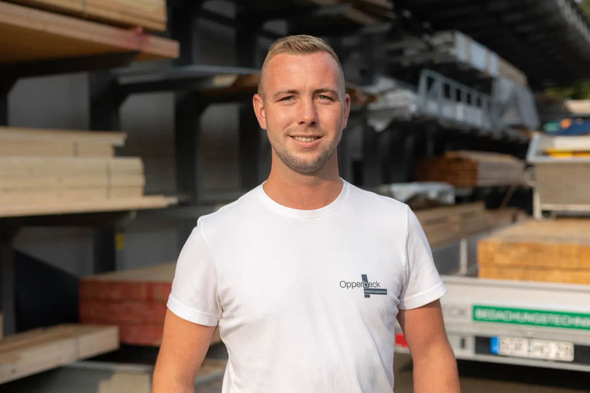 Ein Mann in einem weißen T-Shirt steht vor einem Holzstapel.