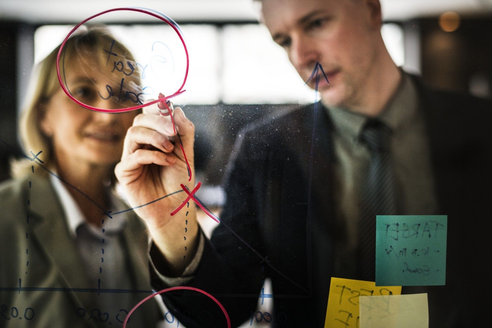 Dos personas intercambian ideas en una pizarra de cristal con notas adhesivas y anotaciones con rotulador.