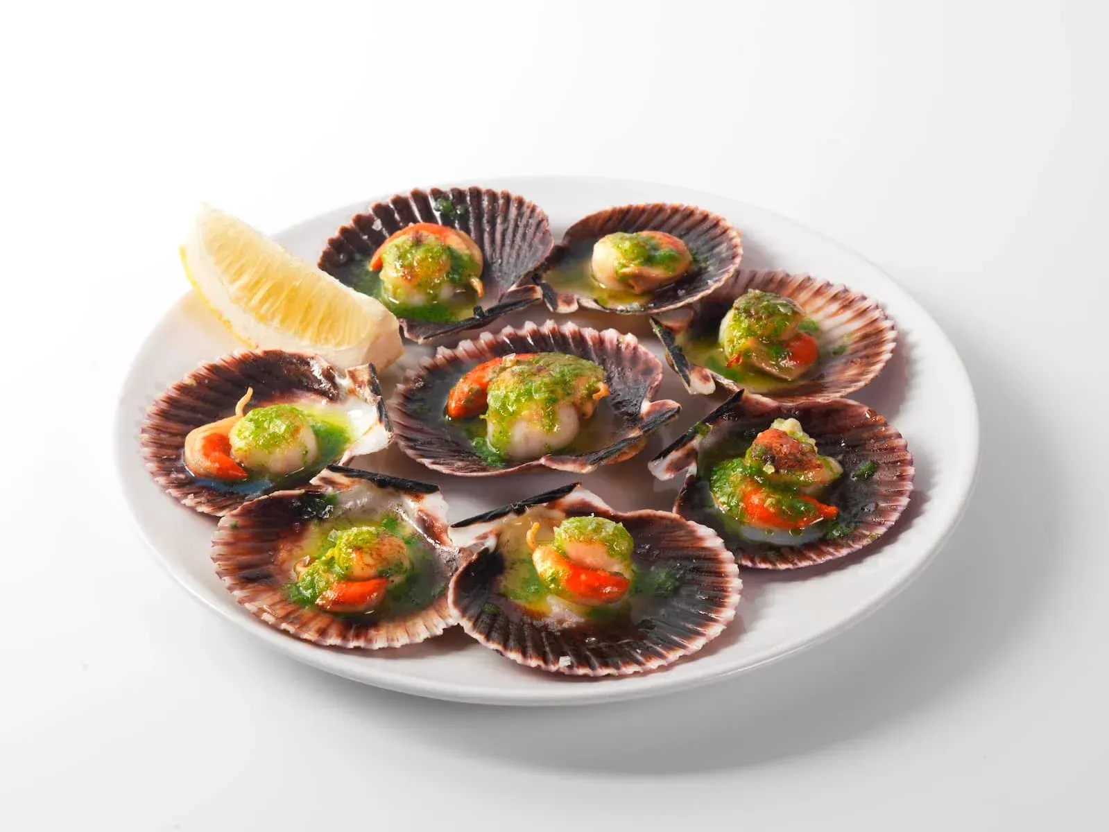 Vieiras selladas en su concha, cubiertas con salsa verde y guarnición roja, servidas en un plato blanco con rodajas de limón.