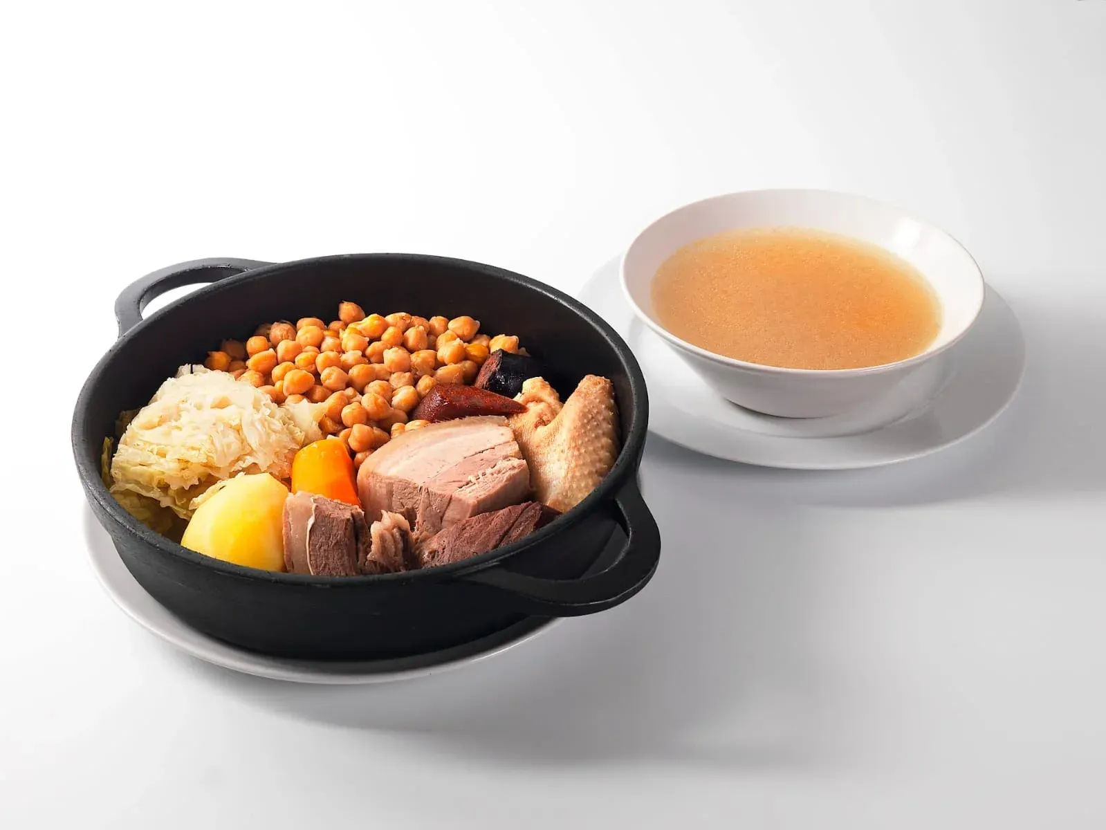 Un tazón negro con carne mixta, papas y garbanzos junto a un tazón de caldo sobre una superficie blanca.