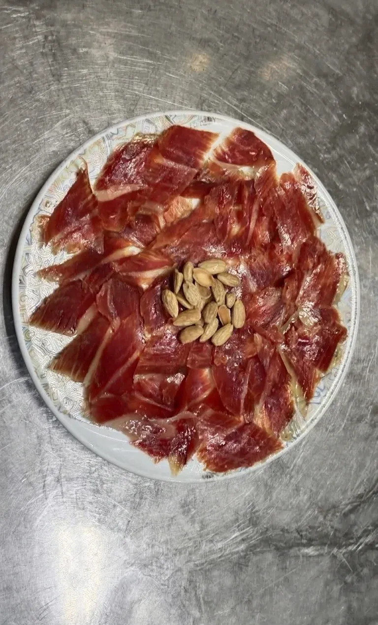 Plato de jamón curado en lonchas finas dispuesto alrededor de un pequeño montón de frutos secos sobre una encimera de acero inoxidable.