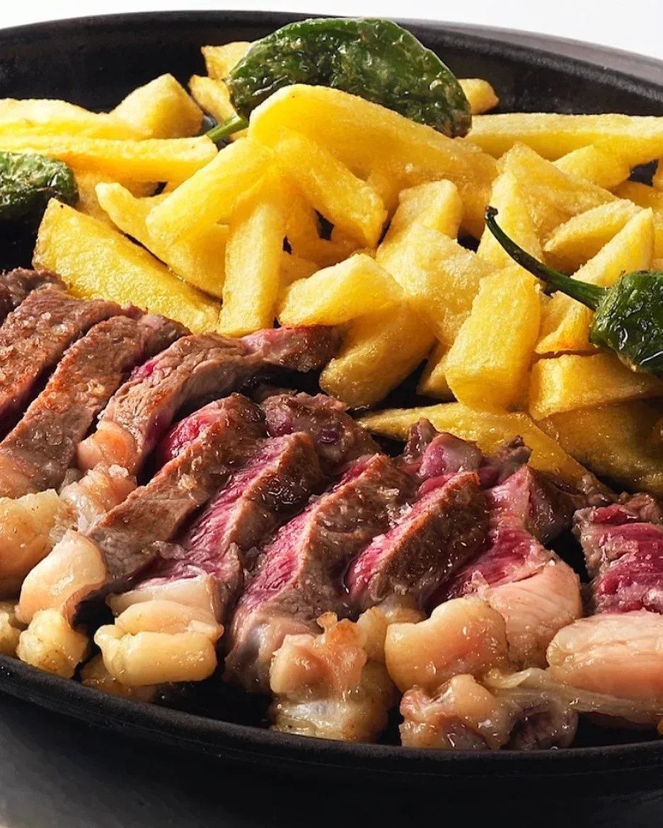 Filete en rodajas con patatas fritas y verduras servido en una sartén negra.