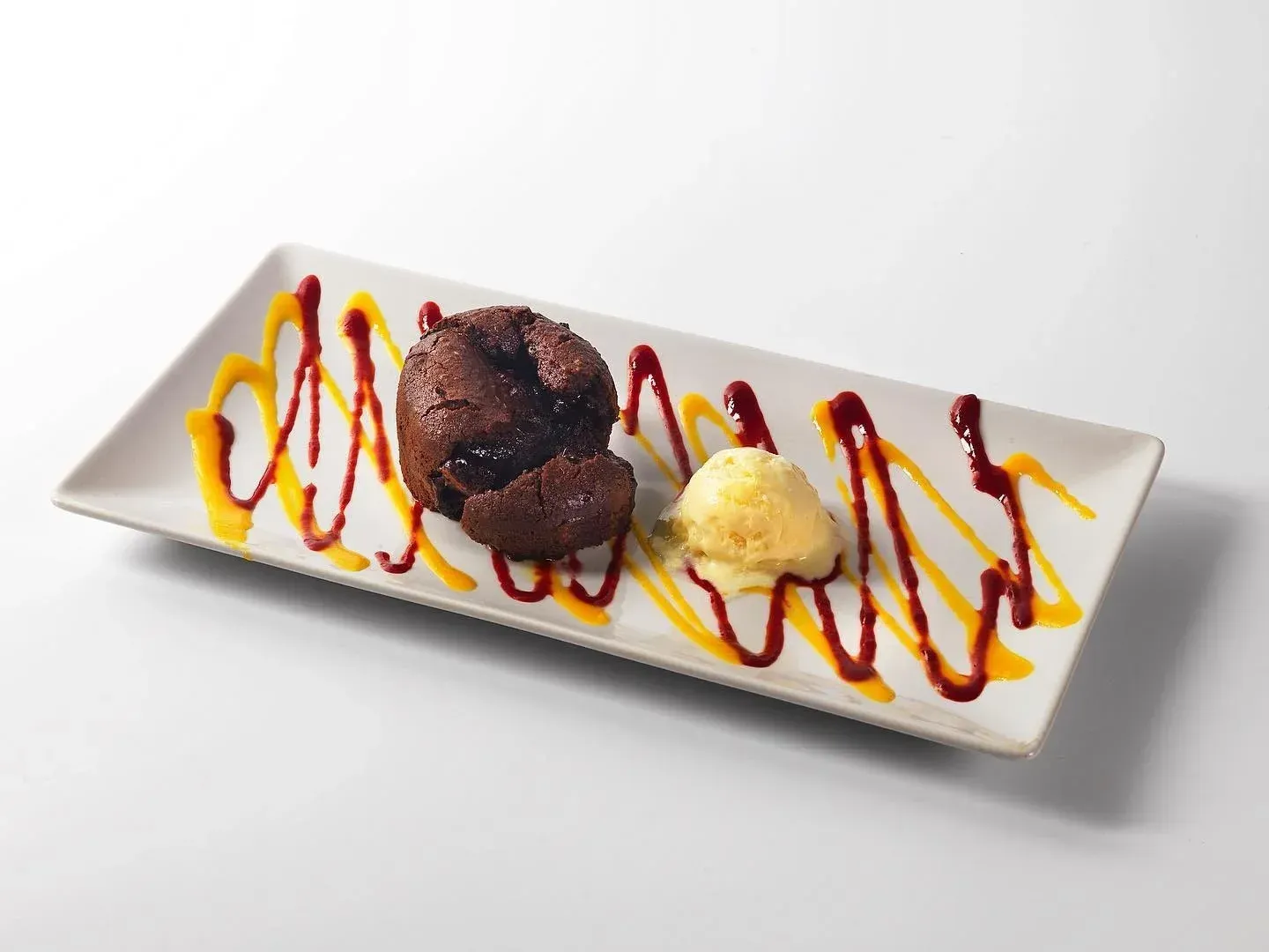 Postre de chocolate con helado de vainilla en un plato blanco, bañado con salsa roja y amarilla.