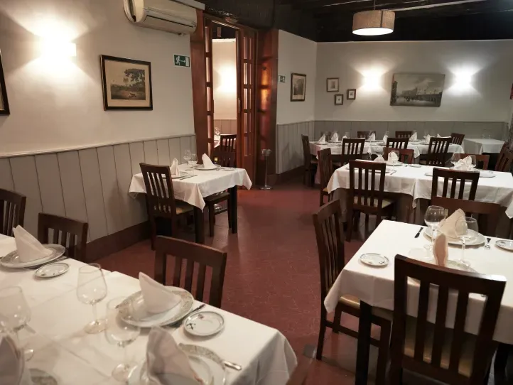 Comedor vacío de un restaurante con mesas puestas, manteles blancos y sillas de madera bajo una iluminación cálida.