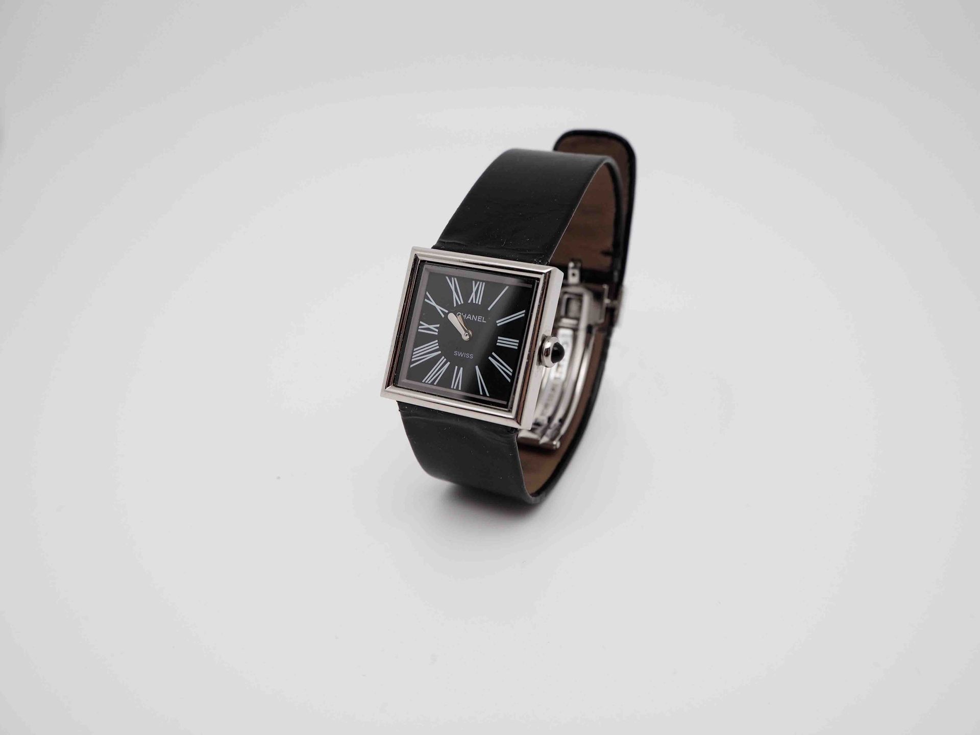 Un reloj cuadrado con una correa de cuero negra está sobre una superficie blanca.