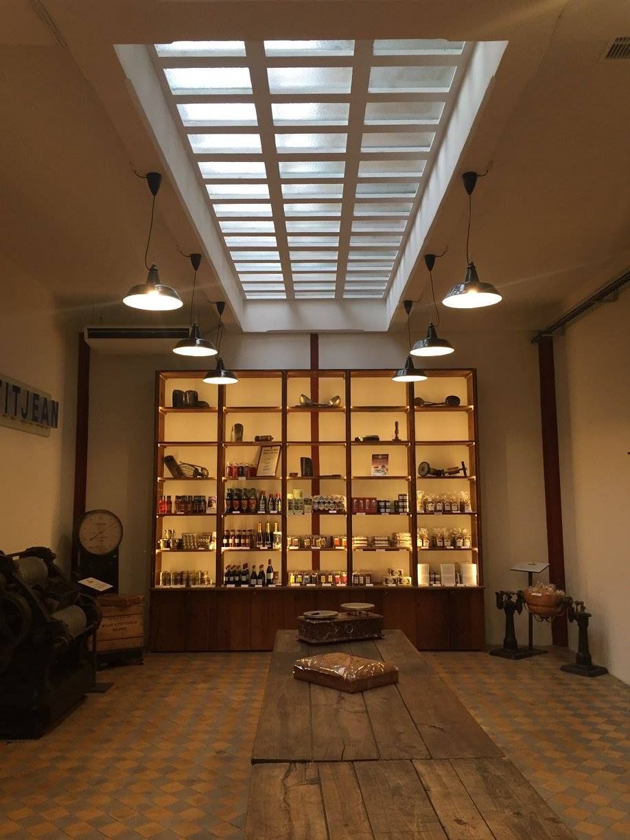 Une pièce rustique avec une longue table en bois, une haute étagère éclairée remplie d'objets et un plafond percé d'un puits de lumière.