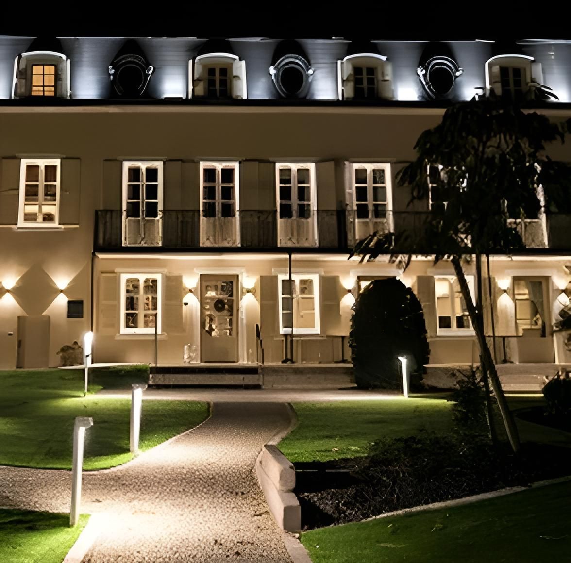 Bâtiment illuminé la nuit, avec un chemin menant à l'entrée, éclairé par des lumières extérieures.