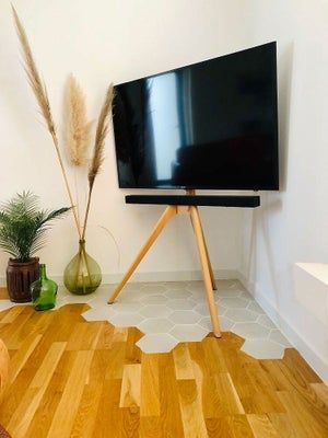 Un téléviseur moderne sur un trépied en bois, adossé à un mur blanc, avec un décor et un sol de formes variées.