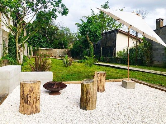 Cour arrière avec gravier, tabourets en rondins, foyer, parasol et pelouse.