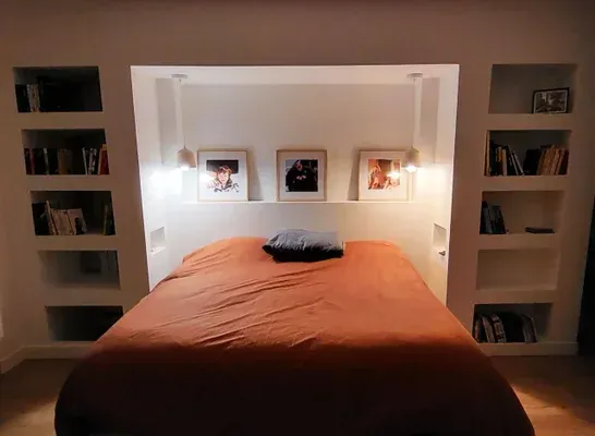 Lit avec linge de lit orange, placé au centre sous des étagères encastrées et des œuvres d'art, avec deux étagères latérales.