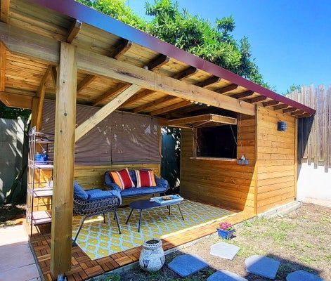 Terrasse extérieure en bois avec sièges, tapis et télévision murale sous un toit partiel.