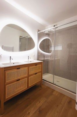 Salle de bains avec meuble-lavabo en bois, miroirs ovales avec éclairage LED et douche en verre.