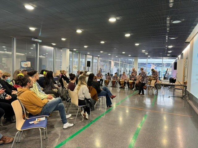 Des personnes assistent à un événement, certaines assises, d'autres jouant du tambour. Un écran de projection est installé sur le côté.