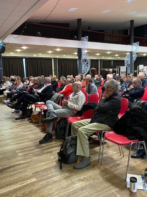 Public assis sur des chaises rouges lors d'une conférence. Sol en bois, murs sombres, décorations.