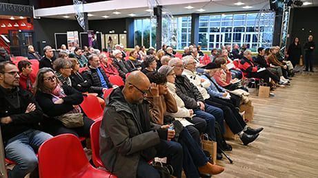 Le public, assis sur des chaises rouges, écoute attentivement dans une grande salle avec une scène.