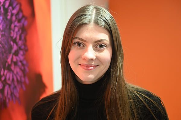 Femme aux longs cheveux bruns, portant un col roulé noir, souriant devant un mur orange.