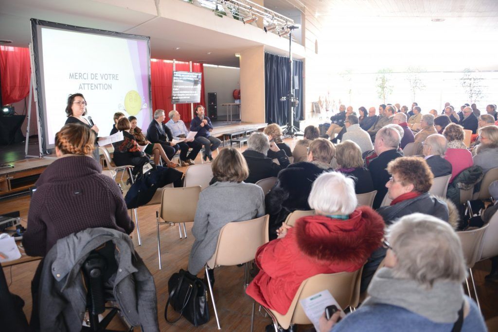Public présent à l'événement ; orateurs sur scène. Grande salle ; lumière du soleil. Personnes assises, certaines écoutant, d'autres regardant la scène.
