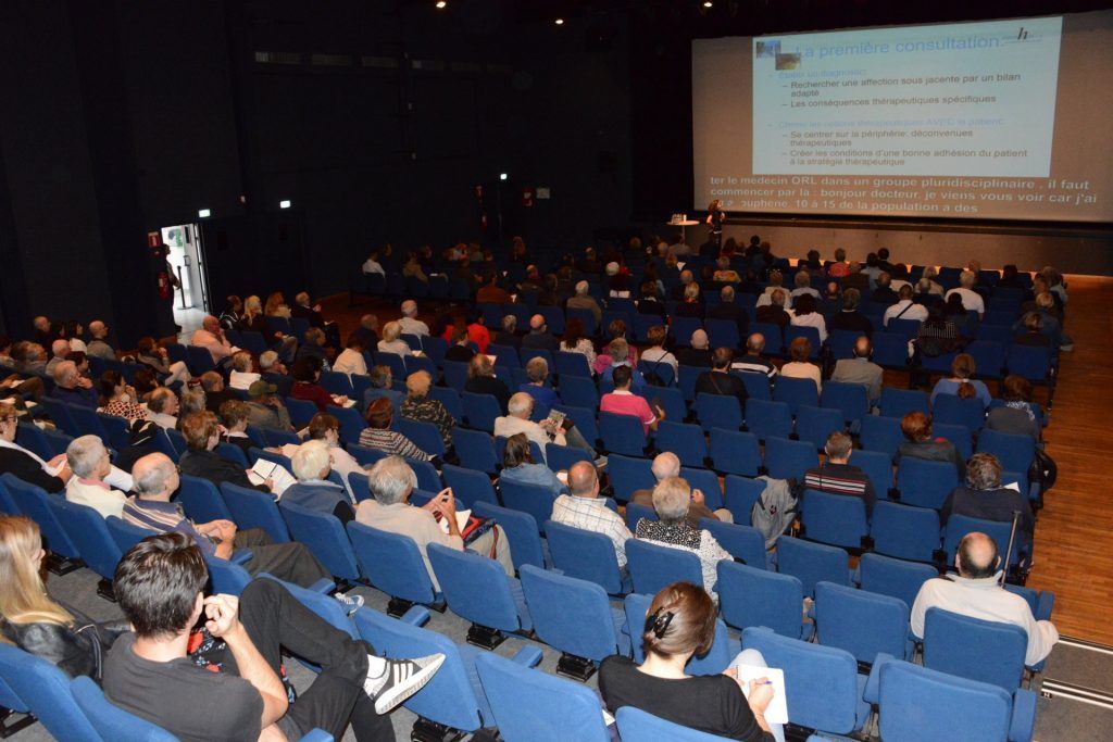 Un public assis dans un auditorium aux sièges bleus regarde une présentation sur un écran.