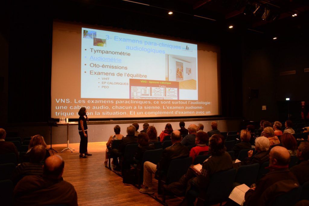 Un présentateur se tient devant un écran diffusant une présentation dans un auditorium rempli de spectateurs.