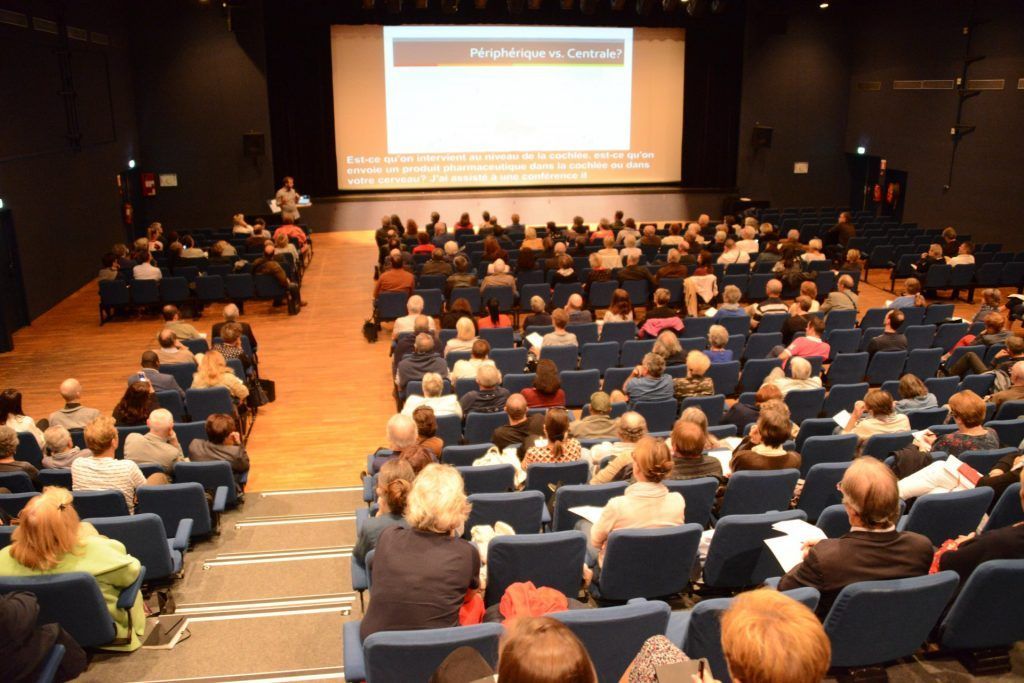 Public dans un théâtre avec un présentateur sur scène et un écran affichant du texte.