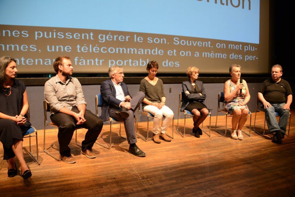 Sept personnes assises sur scène, formant ce qui semble être une table ronde, avec du texte affiché sur un écran derrière elles.