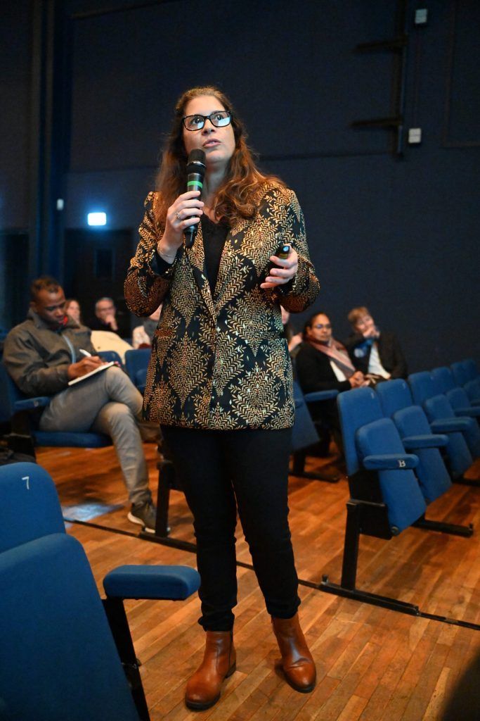 Une femme portant des lunettes parle dans un microphone sur scène, le public est assis sur des chaises bleues.