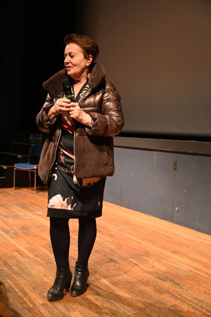 Femme sur scène, vêtue d'un manteau et d'une robe marron, tenant un objet, tournée vers la gauche. Sol en bois, fond sombre.
