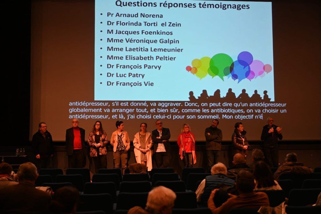 Table ronde sur scène, avec projection sur écran de la liste des intervenants. Public assis dans un théâtre.