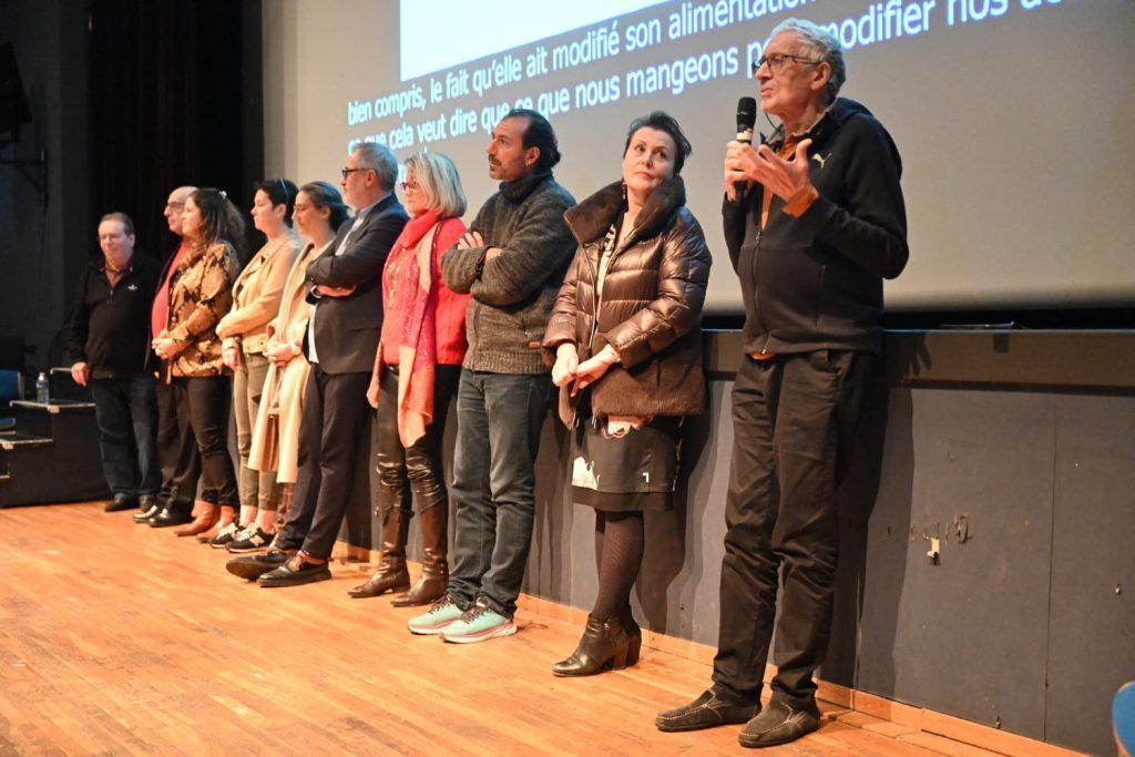 Un groupe de personnes se tient sur scène ; certaines écoutent, une autre parle dans un microphone. Fond sombre avec du texte.
