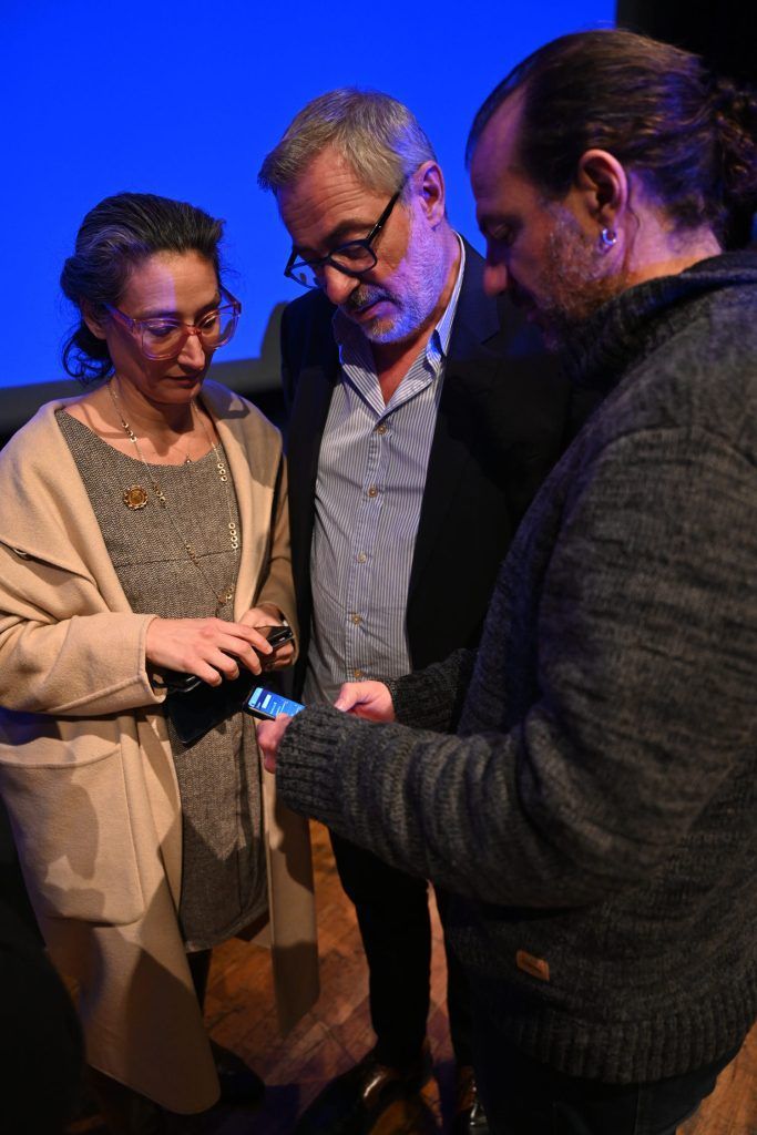 Trois personnes observent un objet ; deux se penchent en avant, la troisième le tient. Ambiance sombre, fond bleu.