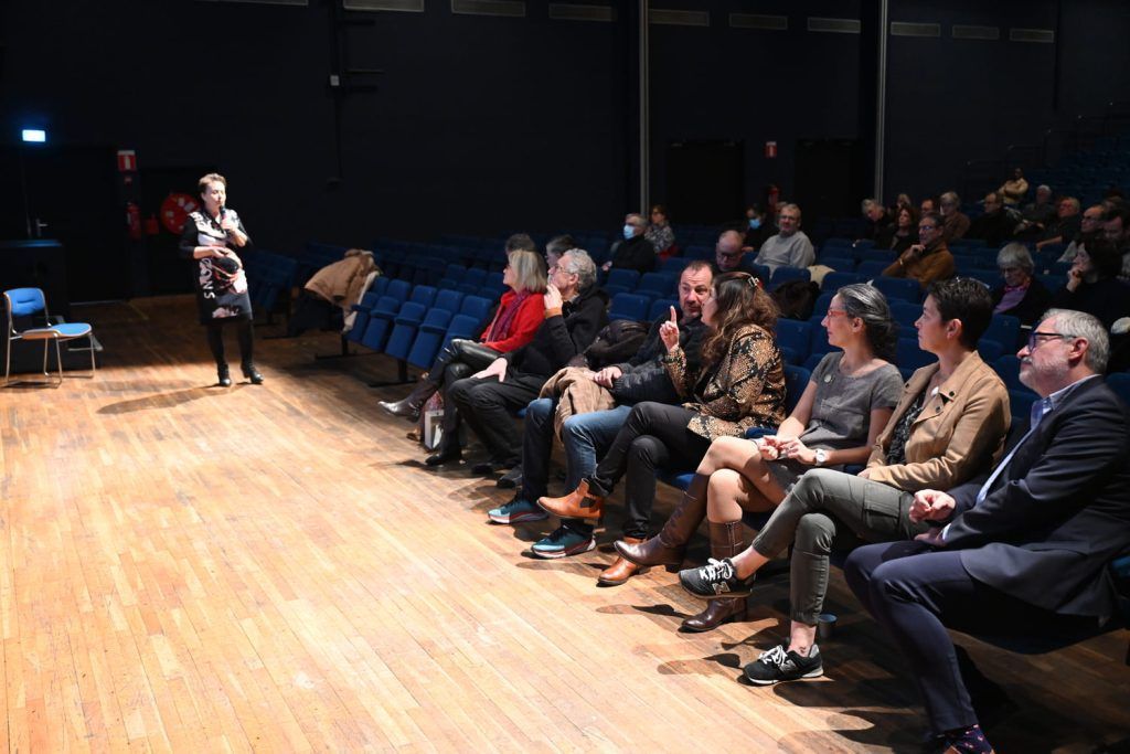 Une personne prend la parole sur scène dans un théâtre, s'adressant à un public assis en rangs.