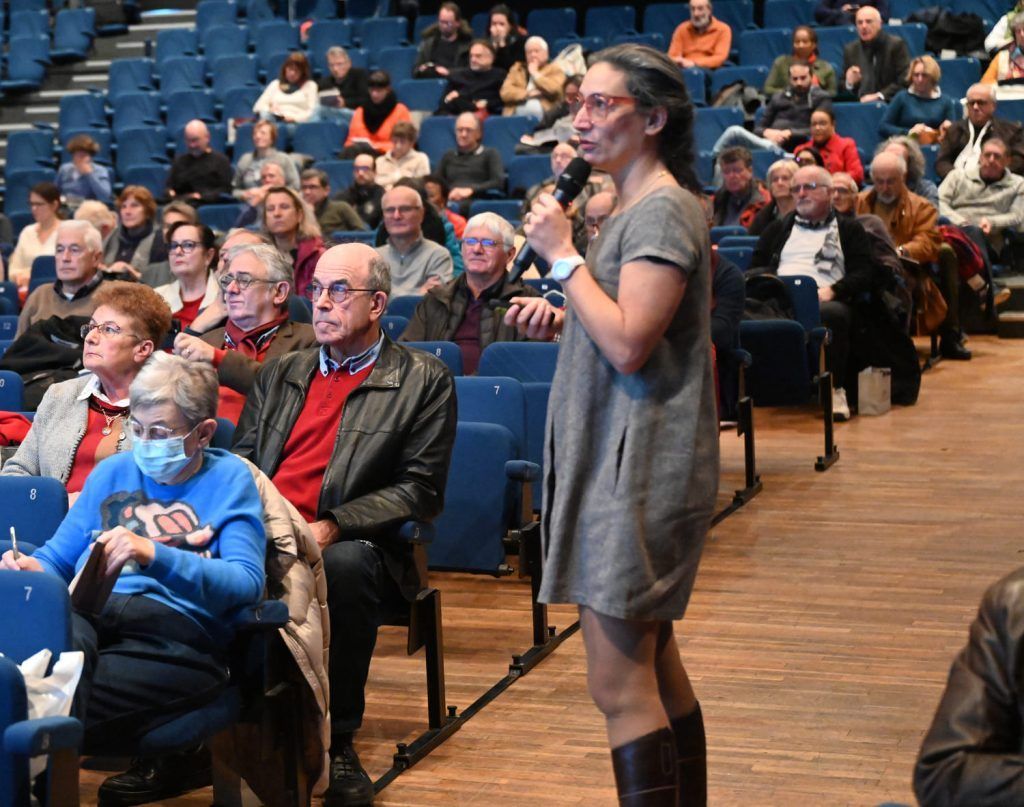 Une femme prend la parole au micro lors d'un événement, debout devant un public assis dans un théâtre.