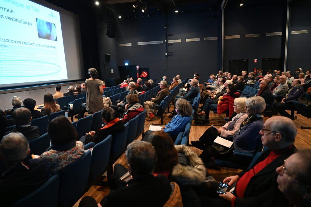 Dans un auditorium plongé dans l'obscurité, le public écoute une présentation sur un écran. Un présentateur se tient devant.