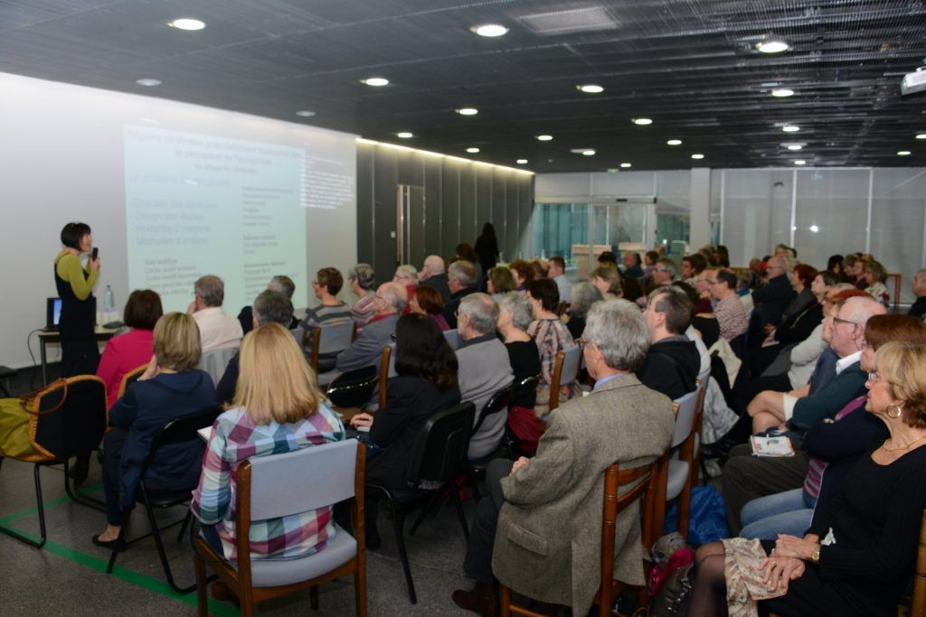 Une femme fait une présentation dans une salle devant un public assis sur des chaises ; un écran affiche la présentation.