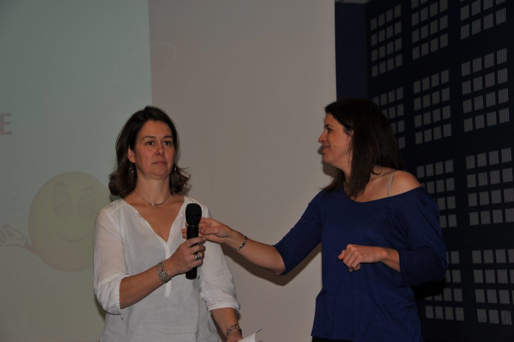 Deux femmes sur scène ; l’une parle dans un micro, l’autre gesticule. Mur blanc et fond carrelé bleu.