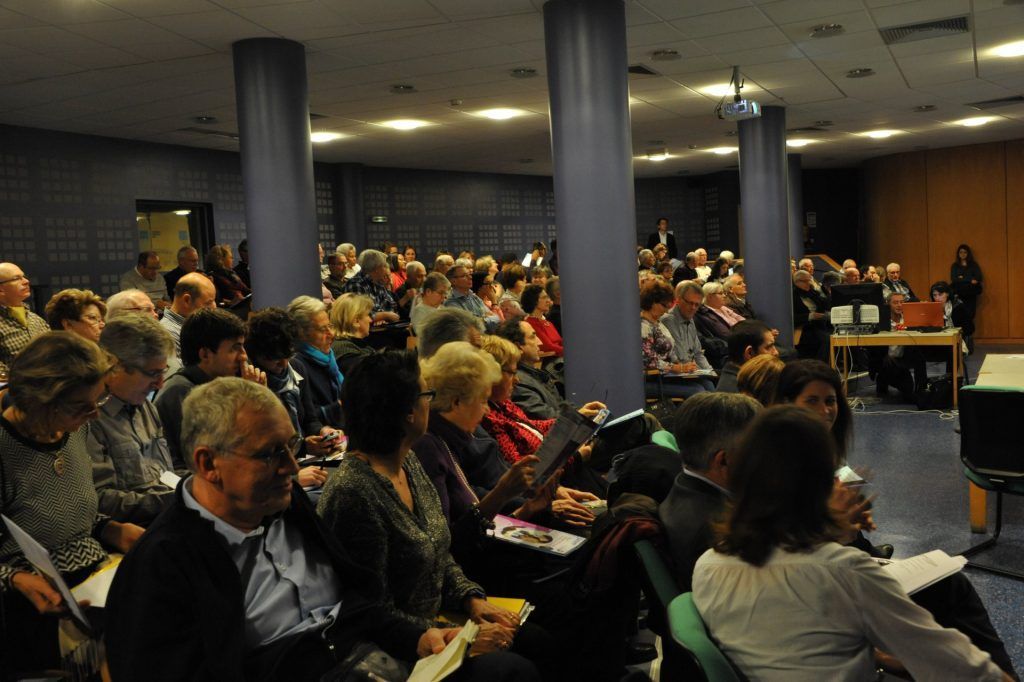 Un public est assis dans une grande salle, assistant à une présentation. On distingue des piliers bleus, un écran blanc et un éclairage tamisé.