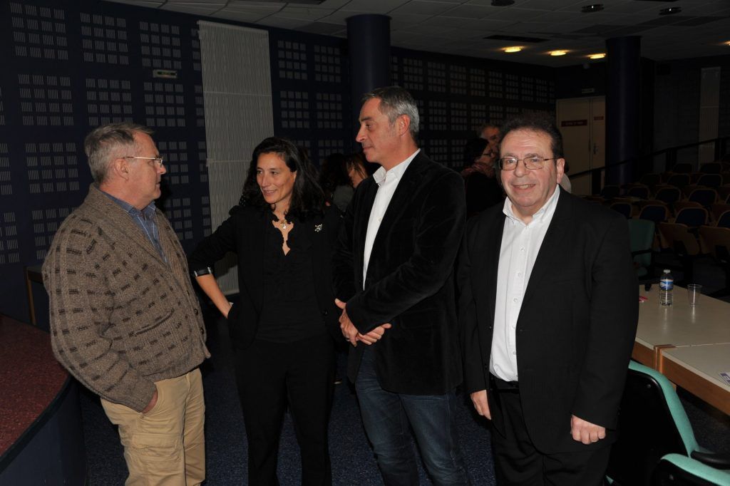 Quatre personnes discutent dans une pièce. Une femme vêtue de noir se tient entre deux hommes, l'un portant une veste et l'autre un pull. Un autre homme se tient à l'écart.