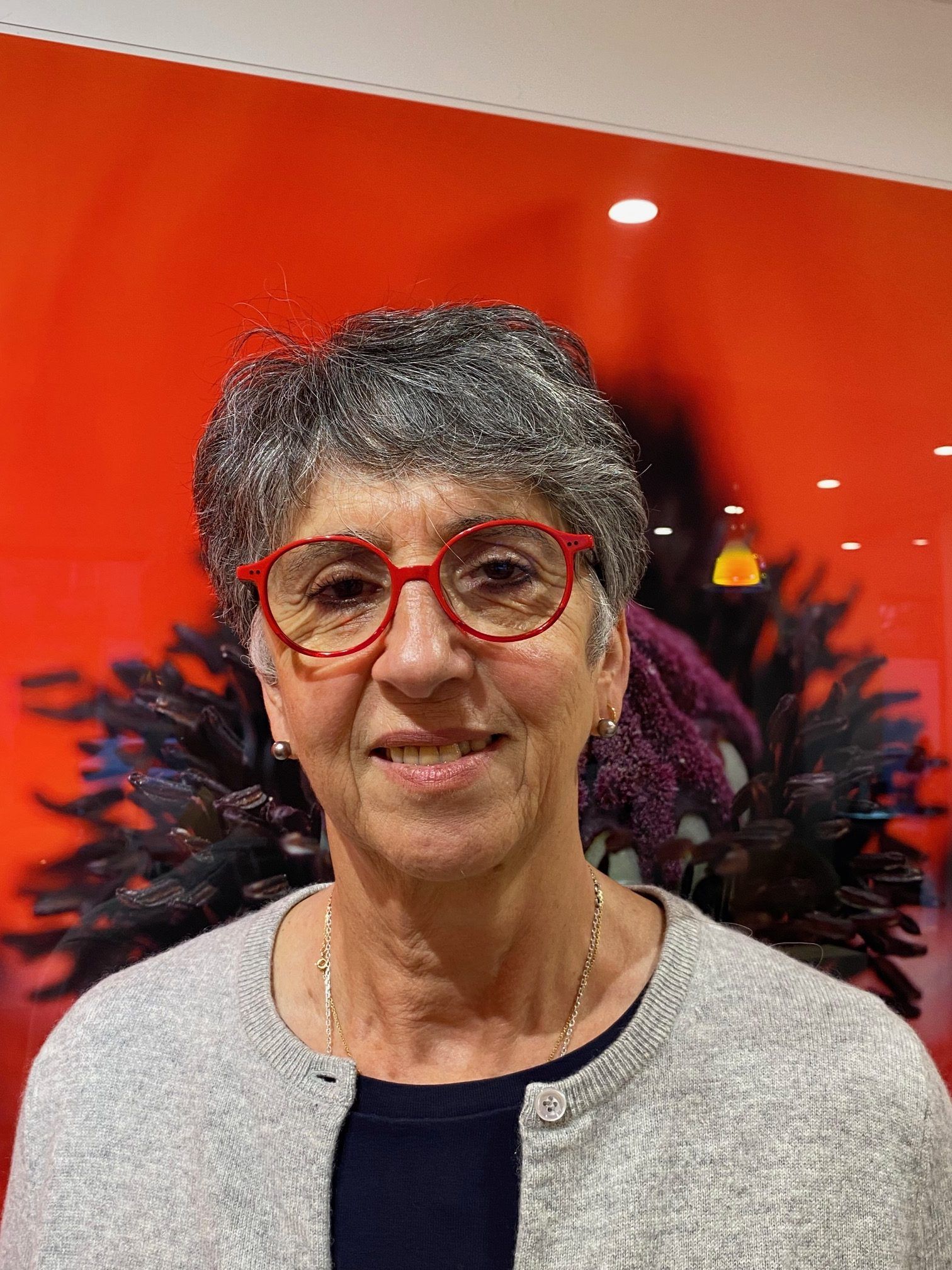 Femme aux cheveux gris courts et aux lunettes rouges, vêtue d'un cardigan gris, souriante, devant un mur rouge orné de motifs floraux.