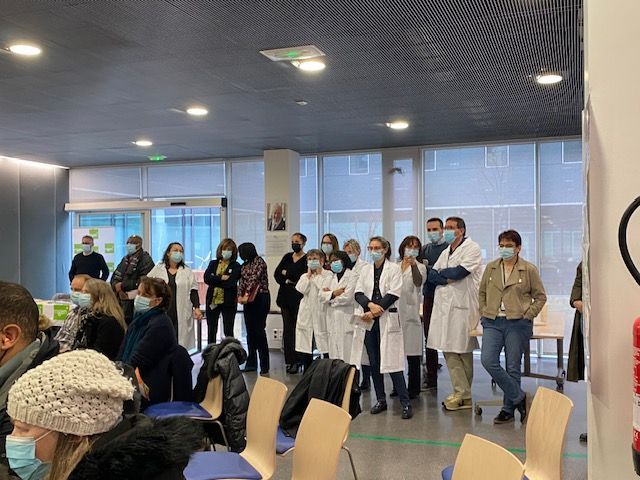 Des personnes, dont du personnel médical en blouse blanche, se tiennent dans une pièce, probablement un établissement médical, portant des masques.