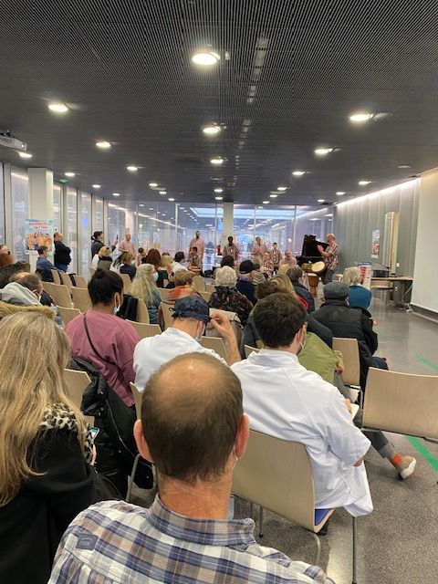 Le public, assis, regarde les artistes sur scène dans un espace intérieur bien éclairé par de grandes fenêtres.