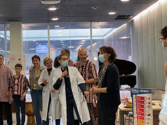 Un groupe de personnes en blouse blanche et masques ; l'une d'elles parle dans un microphone. À l'intérieur, près d'une fenêtre et d'un piano.