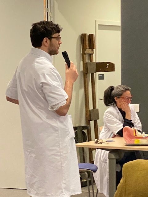 Un homme en blouse blanche parle dans un microphone. Une femme, également vêtue d'une blouse blanche, écoute à une table. À l'intérieur.