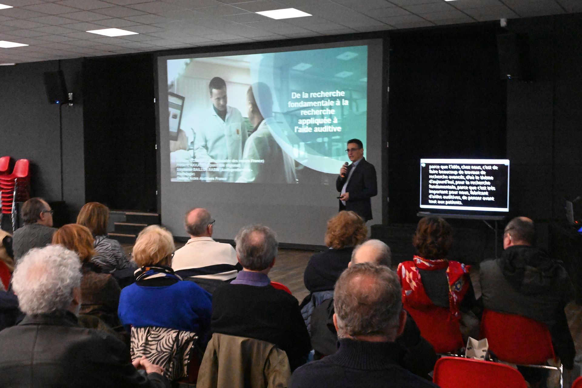 Le public regarde un intervenant donner une conférence dans une salle de réunion, avec une diapositive projetée et un écran.