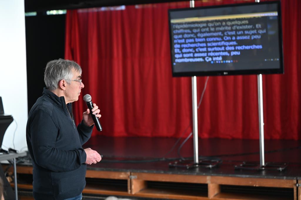 Un homme parle dans un microphone sur scène, devant un écran affichant du texte. Un rideau rouge se trouve en arrière-plan.
