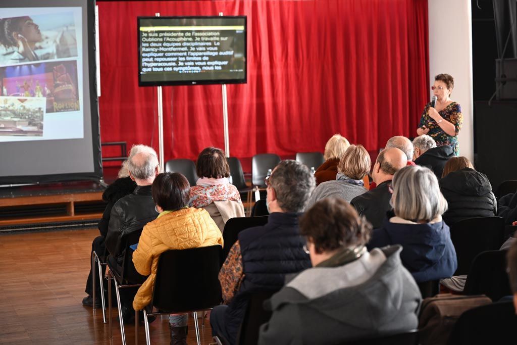 Une femme s'adresse à un public assis dans une salle aux rideaux rouges et devant un écran affichant du texte et des images.