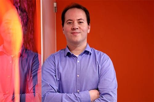 Un homme en chemise bleue se tient devant un mur orange, les bras croisés.