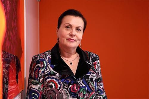 Femme en veste à motifs, appuyée contre un mur orange. Souriante, cheveux courts et foncés.